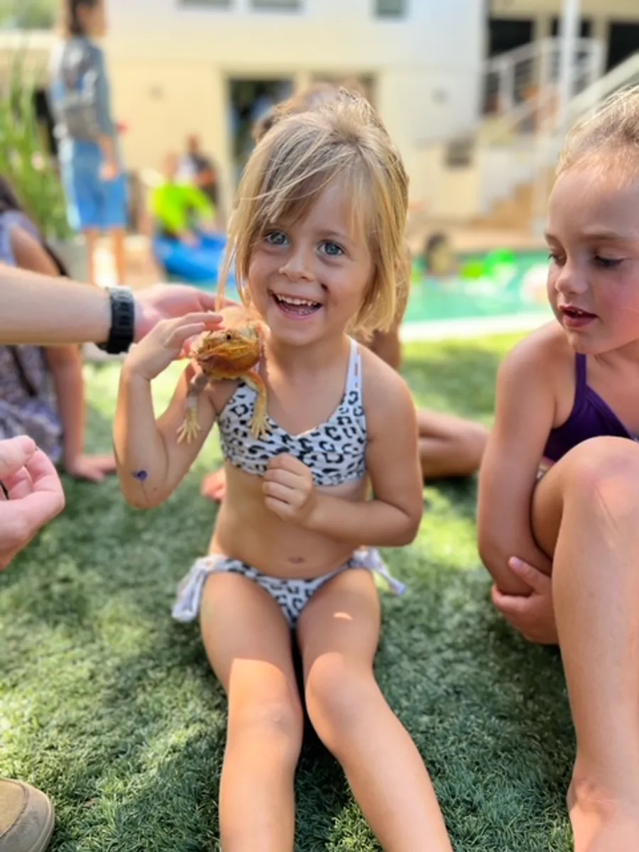 Little girl grinning with a bearded dragon perched on her shoulder at a pool party