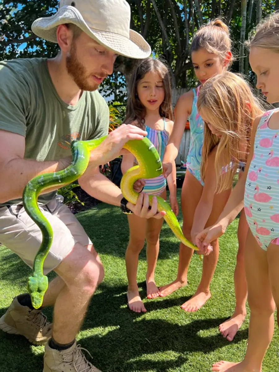 Handler showing a bright green tree python to girls in swimsuits at a backyard pool party